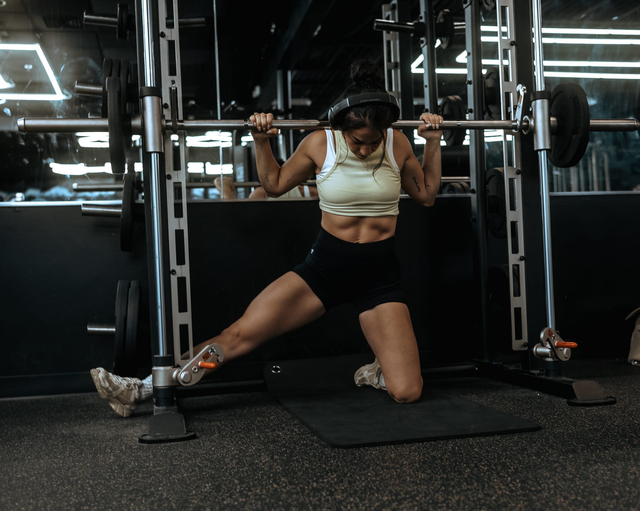 Brooke kneeling under a barbell on a Smith machine, mid-warmup, dark moody gym